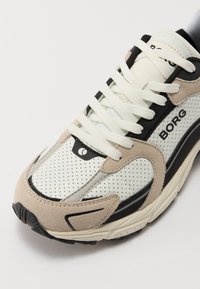 Αθλητικό παπούτσι με λευκό διάτρητο άνω μέρος, μπεζ σουέτ και μαύρες λεπτομέρειες. Μαλακή σόλα με κορδόνια και λογότυπο "BORG" στο πλάι.