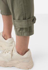 Przycięte zielone spodnie robocze z mankietami i detalem guzikowym, w zestawieniu z masywnymi, off-white sneakerami z teksturowanego materiału.