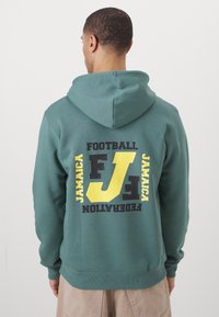 Grön hoodieför med känguruficka och justerbar dragsko på huvan. Stora kontrasterande gula och svarta texter på ryggen, med texten "Football Jamaica Federation."