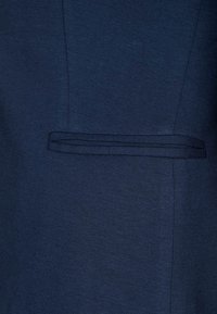 Tmavě modrá tkanina s hladkou texturou a jemným detailem kapsy na levé straně, naznačující střih na míru.