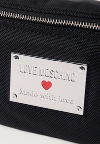 Črna teksturirana torba z zlatim kovinskim znakom, na katerem je vgravirano "LOVE MOSCHINO," rdeče srce in "Narejeno z ljubeznijo." Zadrga za zapiranje.