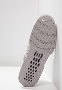 Nike sko sål i lysegrå med en hexagonal slidbane og ventilationshuller. Glat tekstur med en flad profil, vippet mod en væg.