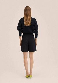 Femme aux longs cheveux portant un haut noir à manches longues et un short noir, se tenant pieds nus sur un fond beige en tournant le dos.