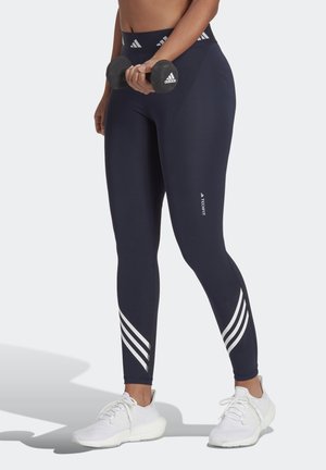 Leggings sportivi blu navy con accenti a strisce bianche, realizzati in tessuto elastico, abbinati a sneakers bianche e con un manubrio in mano.