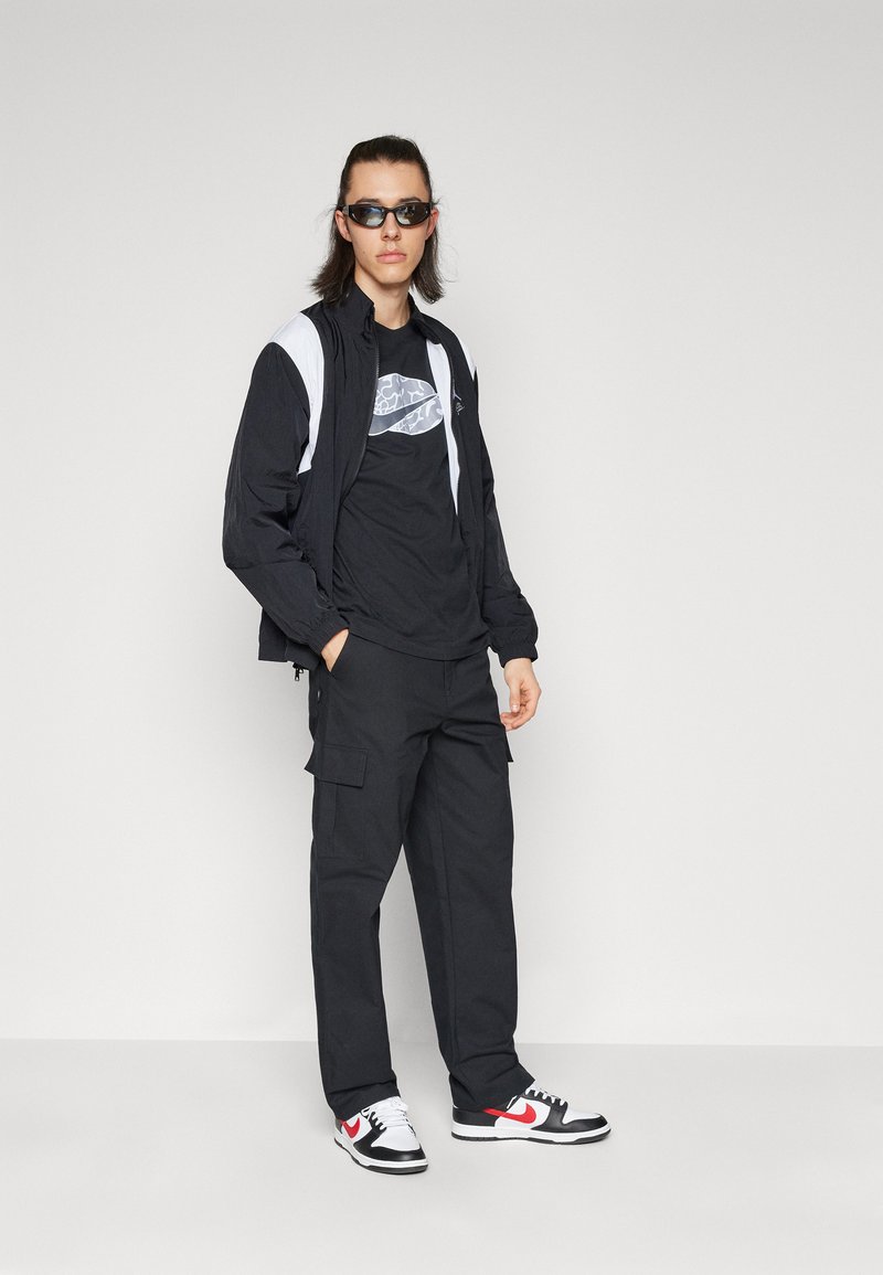 Coupe-vent noir avec des accents blancs, t-shirt graphique noir, pantalon cargo noir et baskets noir/rouge. Les accessoires comprennent des lunettes de soleil sombres.