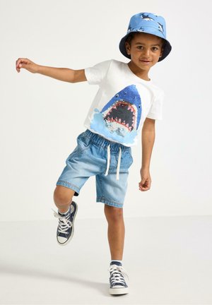 Dreng, der balancerer på ét ben iført en t-shirt med hajmotiv, denimshorts, blå sneakers og en blå bøllehat med hajtryk.