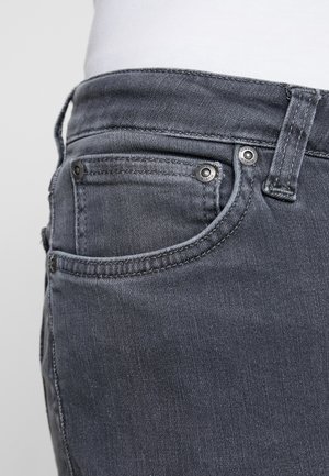 Gros plan sur la poche d'un jean en denim noir et la passants de ceinture avec des coutures visibles et des rivets métalliques sur une personne portant une chemise blanche.
