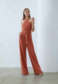 Roestkleurige jumpsuit met een mouwloos ontwerp, voorzien van een strik taille en brede pijpen. Glad stof met minimale details.