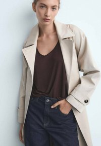 Trench-coat beige avec un col, porté sur un haut marron à col en V et un jean en denim foncé. Dispose de poignets boutonnés et d'une coupe décontractée.