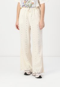 Pantalons légers couleur beige, amples, avec des motifs en œillet et un ourlet festonné. Fabriqués dans un tissu texturé, ils disposent d'une taille élastique et d'un cordon de serrage.