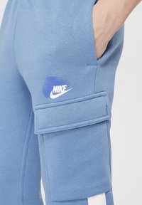 Modré tepláky z měkké látky s velkou nákladní kapsou, bílým logem Nike a kontrastními bílými pruhy na bocích.