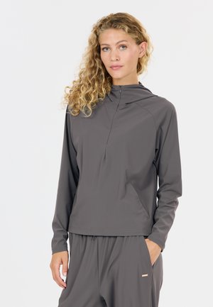 Frau mit lockigem blondem Haar, die ein graues Kapuzen-Longsleeve und passende graue Hosen trägt, mit den Händen in den Taschen.