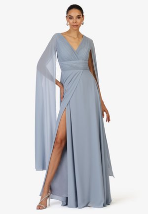 Frau in einem hellblauen Kleid mit plissiertem Oberteil, Cape-Ärmeln und hohem Schlitz, trägt silberne High Heels, steht vor weißem Hintergrund.