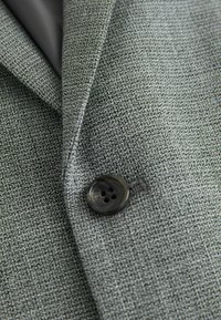 Tela de blazer gris texturizada con un primer plano de un solo botón negro de cuatro agujeros y solapa doblada.