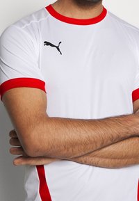 Hvid atletisk skjorte med røde detaljer ved kraven og ærmerne. Har meshpaneler på skuldrene og et sort Puma-logo på brystet.