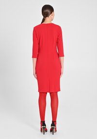 Abito rosso con maniche a tre quarti, design aderente, zip sul retro e spacco posteriore, abbinato a collant rossi e scarpe con tacco nere.
