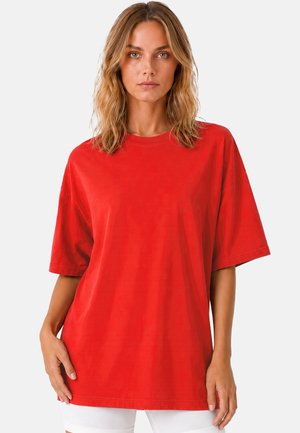 Femme aux cheveux blonds ondulés portant une chemise ample rouge vif à manches courtes et un short blanc, debout devant un fond blanc.