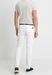 Mężczyzna w białych spodniach, jasnoszarej koszulce, czarnym pasku i czarnych butach stoi tyłem na białym tle.