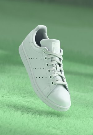 Baskets blanches Adidas Stan Smith avec lacets, côtés perforés et logo sur la languette, flottant au-dessus d'une surface texturée verte douce.