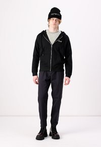 Felpa nera con zip e logo, t-shirt grigia sotto, pantaloni scuri e stivali neri con lacci. Indossa un berretto nero. Look casual.