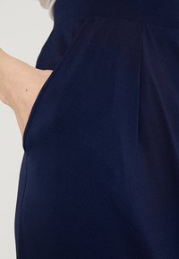 Námořnické modré kalhoty s boční kapsou a plisovaným designem. Tkanina má hladkou, jemnou texturu, která působí elegantně.