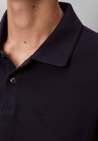 Közeli kép egy sötét tengerészkék galléros, kétgombos pólót viselő személyről, amelyen a mellrészen egy kis hímzett "cK" logó látható.