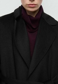 Czarny wełniany płaszcz z szerokim kołnierzem, noszony na ciemnofioletowym golfie. Miękka faktura, swobodny krój, zapinany paskiem w talii.