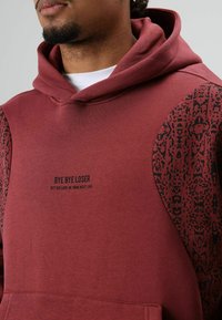 Sweat à capuche bordeaux avec capuche à cordon. Texte noir « BYE BYE LOSER » sur le devant. Accents noirs à motifs sur les manches. Texture douce.