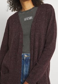 Person som bär en mörk vinröd fluffig cardigan över en olivgrön Afends-skjorta och blå Lee-jeans, beskuren från nacke till höfter.