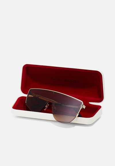 Ochelari de soare maro cu un cadru metalic elegant, având un design cu lentile plate. Aceștia sunt prezentați într-un etui alb cu un interior din catifea roșie.