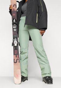 Ski-uitrusting met een zwarte waterdichte jas met neongele accenten, mintgroene broek en zwarte skischoenen, terwijl hij een ski met bindingen vasthoudt.