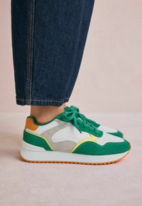Adidași verzi și albi care combină materiale din suede și plasă, cu accente portocalii și galbene. Talpă groasă albă cu un model texturat.