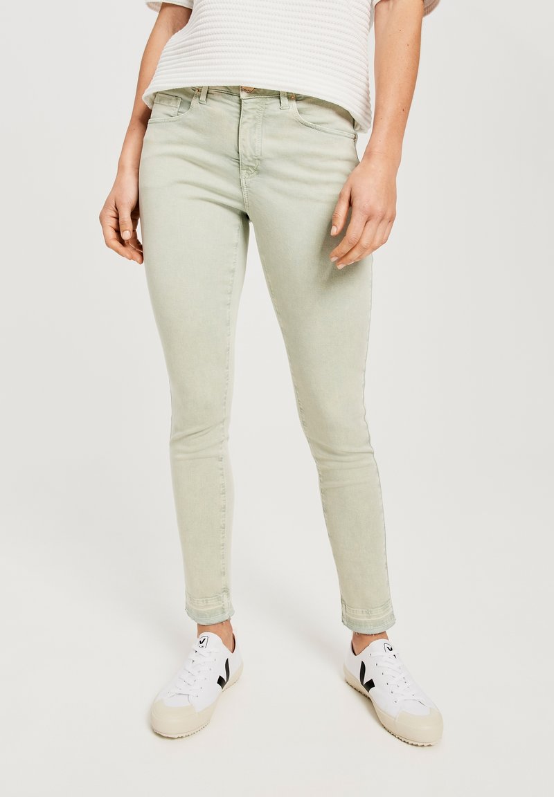 Personne portant un jean slim vert clair, un haut blanc texturé à manches courtes et des baskets blanches à accents noirs, debout devant un fond uni.