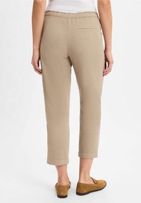 Pantalon beige avec une taille élastique, poignets retroussés et une poche à l'arrière, associé à des chaussures slip-on brun. Tissu lisse et léger.