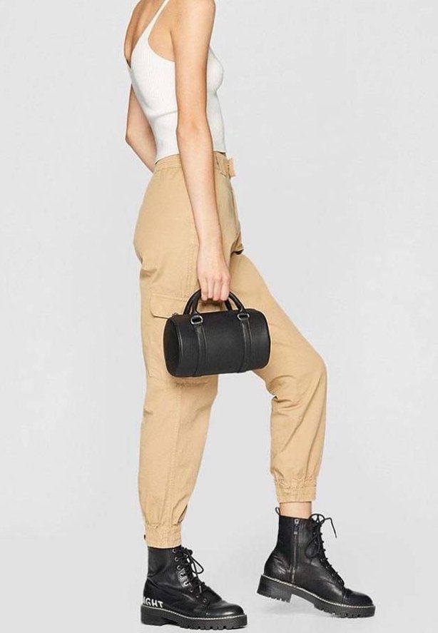 Femme portant un pantalon cargo beige, un débardeur blanc et des bottes noires à lacets, tenant un petit sac à main cylindrique noir à ses côtés.