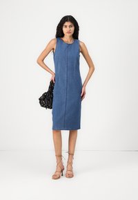 Denim suknelė be rankovių su tiesiu kirpimu, išryškintais siūlų siuvinėjimais priekyje. Modelis laiko juodą gėlėtą rankinę ir vilki sandalus su dirželiais.