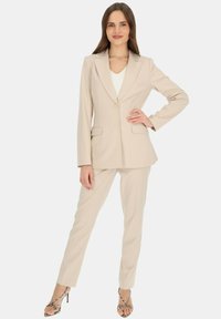 Beżowy, dopasowany garnitur z jednorzędową marynarką posiadającą dwie przednie kieszenie oraz spodnie, wykonany z gładkiego materiału, połączony z szpilkami.