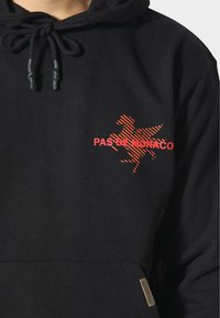 Sudadera negra con bolsillo tipo canguro, capucha con cordón y un gráfico naranja de un caballo con el texto "PAS DE MONACO". Textura suave.