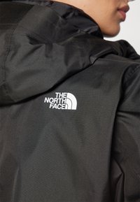 Personne portant une veste à capuche noire avec le logo blanc « The North Face » visible en haut du dos près de l'épaule.