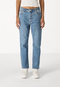 Mujer vestida con jeans de tiro alto y pierna recta en azul claro, zapatillas blancas y una blusa corta blanca texturizada, de pie contra un fondo liso.