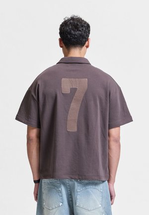 Mężczyzna o krótkich, kręconych włosach, ubrany w brązową koszulkę z krótkim rękawem z dużą cyfrą 7 na plecach oraz jasnoniebieskie szorty dżinsowe.