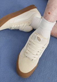 White canvas sneakers with a suede toe cap and brown rubber sole; flat laces, padded tongue, and Vans logo tag.