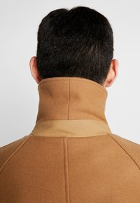 Cheveux courts noirs et oreilles visibles au-dessus du col haut du manteau couleur camel avec un détail en tissu à l'arrière.