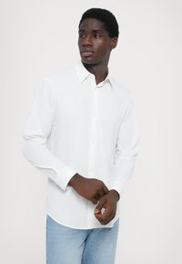 Hvit skjorte med lange ermer og knapper i kragen, glatt tekstur og buet nederkant, sammen med lyseblå denimjeans.