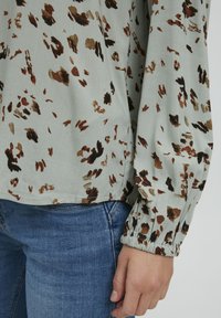 Locker sitzende, hellgrüne Bluse mit braunen und goldenen abstrakten Mustern, elastischen Bündchen und abgerundetem Saum, kombiniert mit blauen Jeans.