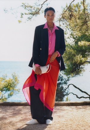 Giovane persona che indossa un blazer nero, camicia rosa, gonna a portafoglio rossa trasparente sopra pantaloni neri, sneakers bianche, tiene una borsa bianca, in piedi all'aperto vicino ad alberi e acqua.