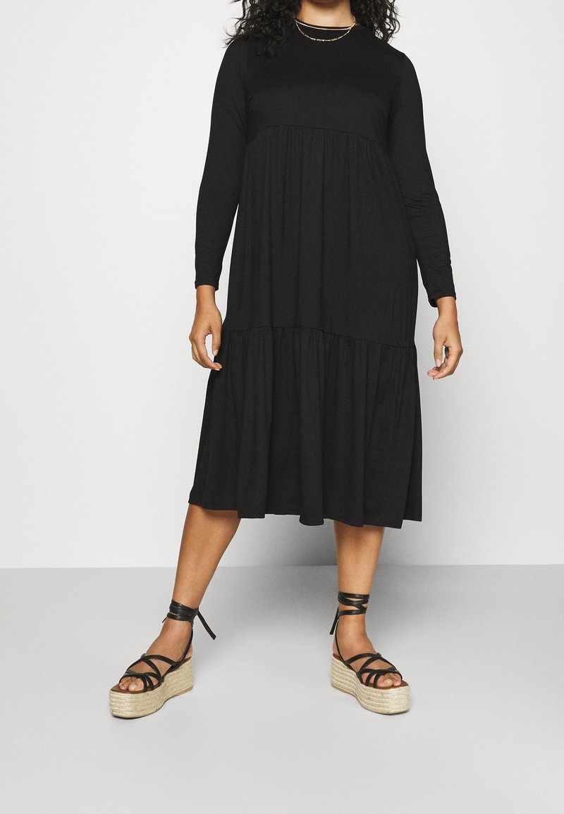 Femme portant une robe midi noire à manches longues avec une jupe à volants et des sandales compensées noires à nouer à la cheville, sur un fond uni.