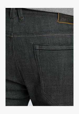 Jean en denim foncé avec une surface texturée, présentant des coutures jaunes contrastantes, une étiquette en cuir et un design de poche arrière standard.