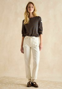 Gestreiftes Langarmhemd in Marineblau und Senfgelb, kombiniert mit hochtaillierten, cremaweißen Hosen. Das Model trägt braune Schuhe und gepunktete Socken.