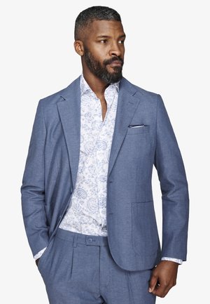 Mężczyzna z krótkimi włosami i brodą, ubrany w niebieski garnitur i białą koszulę w wzory, stoi z jedną ręką w kieszeni, patrząc w bok.